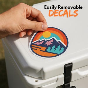 May include: A round sticker with a mountain and sunset design is being applied to a white cooler. The sticker features a mountain range, trees, and a sun, with a rainbow arc. The text "Easily Removable DECALS" is above the sticker.