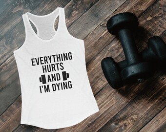May include: White tank top with black text that reads "EVERYTHING HURTS AND I'M DYING" and a dumbbell graphic. A black dumbbell rests on a wooden surface next to the tank top. Fitness apparel.