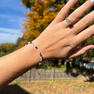 Op de afbeelding: Een close-up van iemands pols met een wit en blauw kralenarmbandje met een zilveren bedel. De persoon draagt ook een zilveren ring met een grote, heldere edelsteen.
