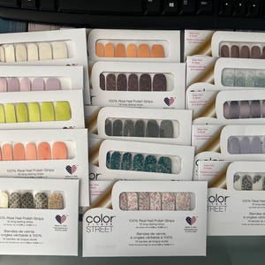 Op de afbeelding: Een verzameling Color Street 100% echte nagellakstrips in verschillende kleuren en ontwerpen. De nagelstrips zijn verpakt in wit karton met een transparant venster. De ontwerpen omvatten effen kleuren, glitter en patronen.