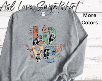 May include: Gray sweatshirt with the word "LOVE" spelled out in American Sign Language, with flowers and a butterfly. The text "ASL Love Sweatshirt" is at the top of the image and "More Colors" is at the top right.