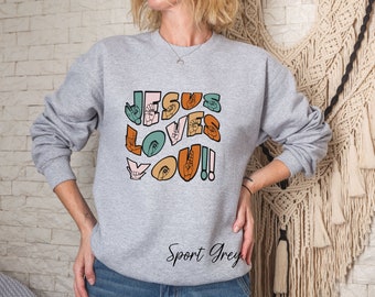 May include: A gray sweatshirt with the text "JESUS LOVES YOU!" in colorful letters formed by hand signs. The text is in a retro style font.