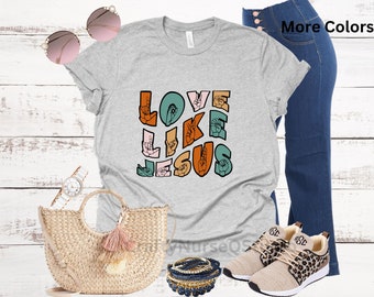 May include: A light gray t-shirt with the words "LOVE LIKE JESUS" in colorful sign language letters. The shirt is paired with blue jeans, a straw tote bag, sunglasses, a watch, bracelets, and leopard print sneakers.