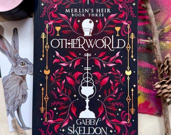 May include: A hardcover book titled "Merlin's Heir Book Three: Otherworld" by Gabby Skeldon. The cover is black with red and gold floral designs and illustrations. A pink candle and a drawing of a rabbit are in the background.