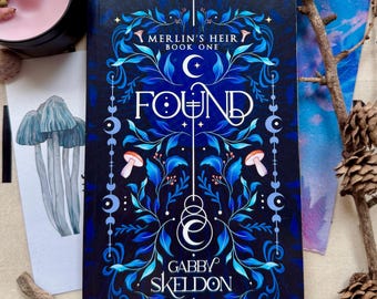 May include: A hardcover book titled "FOUND" by Gabby Skeldon, part of the "Merlin's Heir" series. The cover features a dark blue background with intricate blue and white floral designs, mushrooms, and crescent moons. A candle and artwork are also visible.