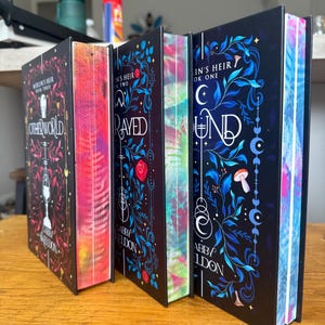 May include: Three hardcover books from the Merlin's Heir series by Gabby Sheldon, titled Otherworld, Betrayed, and Bound. Each black cover features intricate blue, red, and white botanical illustrations, with vibrant, multi-colored sprayed page edges.