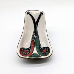 May include: White ceramic pipe rest with two crossed pipes painted in black, green, and red.