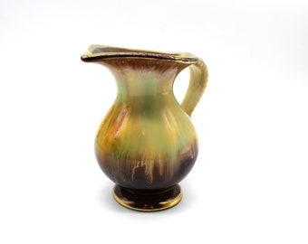 May include: A small, brown, green, and gold ceramic pitcher with a rounded body and a handle. The pitcher has a glazed finish with a mottled pattern.