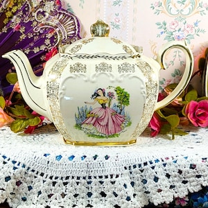 Puede incluir: Tetera vintage de color crema con adornos dorados y una imagen decorativa de una mujer con un vestido rosa. La tetera está sobre un mantel de ganchillo blanco, con una lámpara amarilla y un abanico decorativo en el fondo.