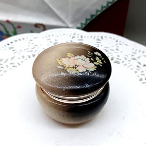 Puede incluir: Una pequeña caja de cerámica redonda de color blanco y negro con un diseño floral en la tapa. La tapa tiene una flor rosa claro con detalles amarillos y blancos.