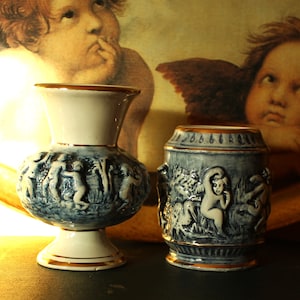 Puede incluir: Dos jarrones de cerámica azul y blanco con ribetes dorados, que presentan diseños en relieve de querubines y otras figuras. Los jarrones se exhiben en una bandeja de madera con una pintura de querubines.