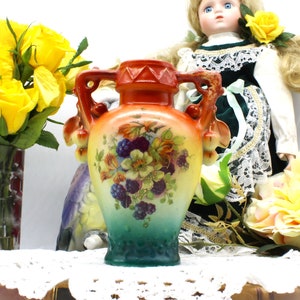 Könnte beinhalten: Eine kleine, handbemalte Porzellanvase mit Blumenmuster. Die Vase hat zwei Henkel und eine Verlaufsglasur, die von Orange nach Grün übergeht. Die Vase ist mit Brombeeren, Blumen und goldenen Akzenten verziert.