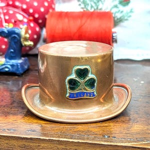 May include: A miniature copper-coloured top hat, featuring a shamrock emblem and the word "IRELAND" in blue. It rests on a wooden surface, with sewing items visible in the background.