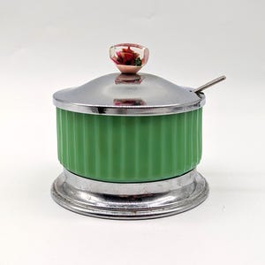 May include: A green, vertically ribbed, cylindrical container with a polished silver-tone metal base and lid. The lid features a small, clear, rounded knob containing a red floral accent. A small silver-tone metal spoon rests inside.