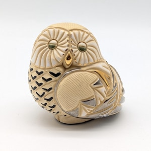 May include: A detailed owl figurine in cream, white, gold, and black. The owl has large, round eyes with gold accents and a textured body. The wings and body feature intricate carved details. The figurine is a decorative item.