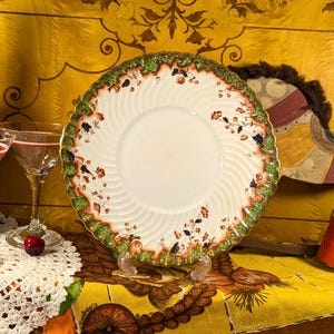 Puede incluir: Naturaleza muerta vintage con un plato decorativo con borde floral, dos copas de martini con bordes rojos y una botella de Cherry Beer. También hay un caballo de juguete de madera y un cojín rojo.