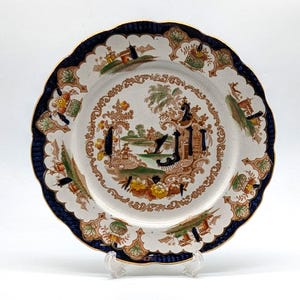 May include: A white ceramic plate with a blue and gold rim. The plate features a detailed floral and landscape design in shades of brown, green, and yellow.