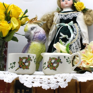 May include: Two white ceramic creamer and sugar containers with a Celtic knot design and shamrock accents. The containers are on a table with a white lace tablecloth and a book.
