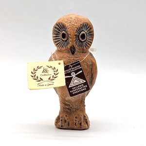 Puede incluir: Una figura de búho marrón con ojos grandes y una superficie texturizada. El búho tiene una etiqueta que dice "Made in Greece" y "Hellenic Handcrafted Creations".
