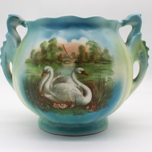 May include: A blue and white ceramic vase with two swan handles. The vase features a painted scene of swans on a lake with trees and a bridge in the background.