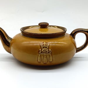 Może przedstawiać: Brązowy ceramiczny czajnik z pozłacanym wykończeniem i herbem korony z napisem "1650" z przodu.
