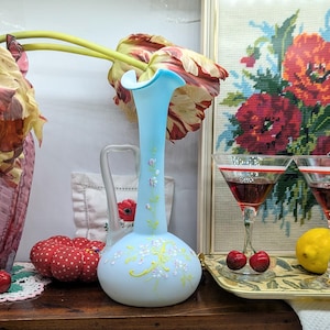 Puede incluir: Un jarrón de cristal azul claro con borde acampanado y detalles florales. El jarrón está junto a un jarrón rosa más grande con un tulipán y un alfiletero de lunares rojos. Dos copas de martini con líquido rojo y cerezas están en una bandeja.