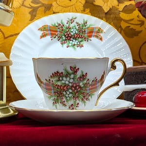 May include: A white teacup, saucer, and plate set with gold trim and a floral and plaid design. A slice of chocolate cake, a cherry, and grapes are also visible. The teacup has a gold handle.