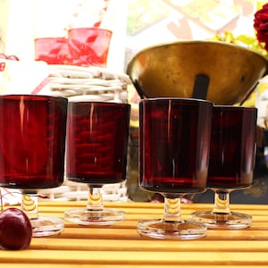 Peut inclure: Quatre verres à liqueur en verre rouge rubis avec des tiges et des bases claires. Les verres sont disposés en rangée sur une surface en bois. Deux cerises rouges sont au premier plan.