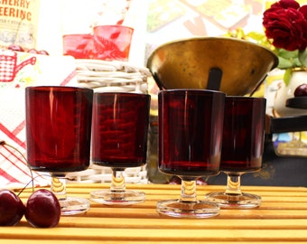 May include: Four ruby red glass shot glasses with clear stems and bases. The glasses are arranged in a row on a wooden surface. Two red cherries are in the foreground.