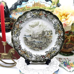May include: A vintage ceramic plate with a green and brown floral border. The centre of the plate features a detailed illustration of a harbour scene with ships and buildings.