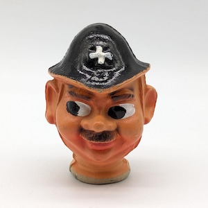 May include: A molded figurine of a head featuring a tan complexion, a dark brown mustache, and large eyes with black pupils. The figure wears a black hat with a white cross emblem on the front. The facial features are painted with pinkish cheeks.