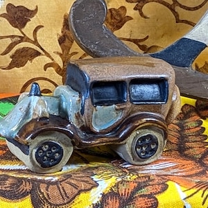Puede incluir: Pequeña figura de coche antiguo de cerámica en tonos marrón, beige y azul. El coche tiene ruedas y ventanas detalladas. El fondo incluye un elemento decorativo de madera y una tela estampada colorida.