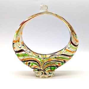 Puede incluir: Una cesta decorativa de vidrio con un asa circular. El vidrio transparente está arremolinado con rayas verdes, naranjas, marrones y negras. La base es de vidrio transparente texturizado. La parte superior del asa tiene un remolino decorativo.