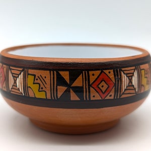 May include: A round terracotta bowl with a white interior and a wide exterior band featuring hand-painted geometric patterns in black, red, and yellow. The design consists of repeating diamonds, triangles, and lines encircling the bowl.