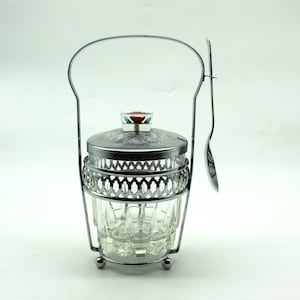 May include: A silver sugar dispenser with a glass jar and a matching spoon. The dispenser has a decorative handle and a floral design on the lid.