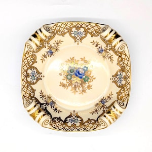 Puede incluir: Cenicero cuadrado crema con diseños de filigrana y florales dorados. El cenicero presenta un ramo floral central en azul, amarillo y verde. El borde está acentuado con rayas doradas y negras.