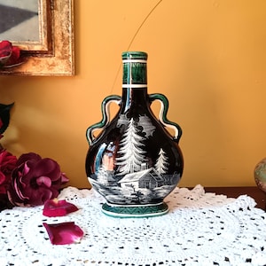 May include: A black and green ceramic bottle with a white scene of a cabin in the woods. The bottle has two handles and a green rim.