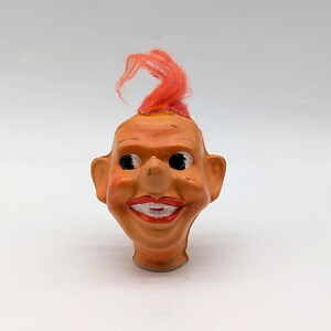 May include: A vintage-style doll head figurine featuring a light orange complexion, prominent ears, and a wide, smiling mouth with painted red lips. The figure has a tuft of bright orange synthetic hair on top and large, dark, round eyes.