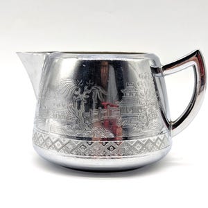May include: A silver-colored creamer with a handle and spout. The creamer features an engraved Asian-inspired landscape design around the body and a geometric pattern at the base. The handle is a curved, open design.