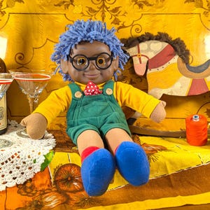 Puede incluir: Una muñeca vintage con cabello azul de hilo, gafas y un mono verde está sentada sobre una superficie amarilla estampada. También hay una botella de licor Cherry Heering, dos copas de cóctel y un caballito de madera. También son visibles una bobina de hilo naranja y un cojín rojo.