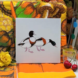 Puede incluir: Una tarjeta blanca con una ilustración en blanco y negro de dos patos. Los patos se miran y tienen la cabeza inclinada. La tarjeta está sobre una tela colorida con una rosa amarilla.