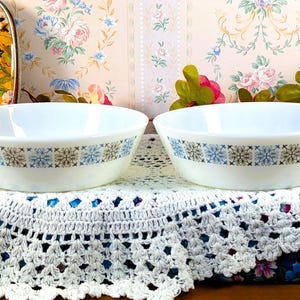 Puede incluir: Dos cuencos de cerámica blanca con un patrón floral azul y marrón alrededor del borde. Los cuencos se exhiben sobre un mantel individual de encaje blanco, con un fondo floral. Los cuencos son probablemente para servir comida.