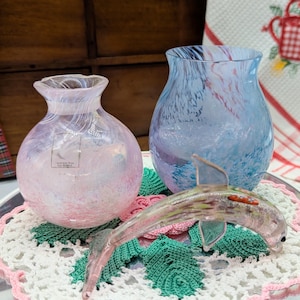 Puede incluir: Dos jarrones decorativos de cristal, uno rosa y otro azul, sobre una bandeja plateada con un tapete de ganchillo. Una figura de delfín de cristal descansa sobre el tapete. Un paño de cocina con un diseño de regadera está en el fondo.