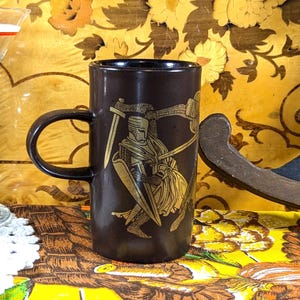 May include: A dark brown ceramic mug with a black handle and a gold illustration of a knight in armour holding a sword and shield. The mug is cylindrical and has a glossy finish. A cherry sits next to a glass.