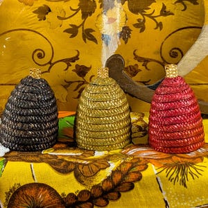 May include: Three decorative, woven beehive-shaped containers in black, gold, and red. Each has a gold-colored top. A cocktail glass with a red rim and a cherry are in the background, along with a yellow floral patterned background.