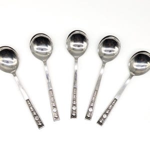 May include: Five silver soup spoons on a white background. Each spoon has a rounded bowl and a long handle with decorative detailing. The spoons are arranged in a fan shape.