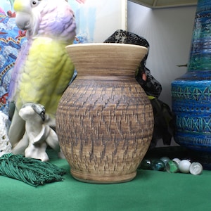 May include: A brown ceramic vase with a textured surface. The vase has a wide mouth and a rounded body. It is sitting on a green surface with a blue and green vase in the background.