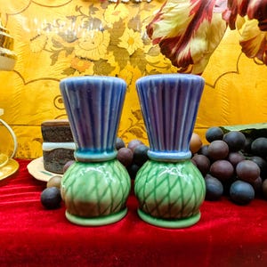 May include: Two decorative ceramic vases with blue tops and green bases, set on a red surface. The vases have a unique shape and are surrounded by grapes and other decorative elements. The background features a yellow floral pattern.