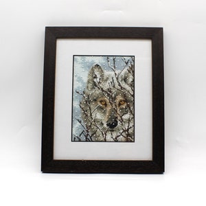 May include: A framed cross-stitch embroidery depicting a wolf's face peering through a snowy forest scene. The wolf has gray fur and piercing yellow eyes.