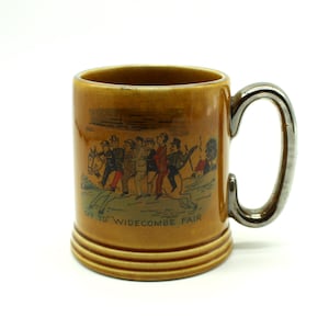 May include: A brown ceramic mug with a silver handle. The mug has a cartoon illustration of people on horseback with the text "Off to Widecombe Fair".